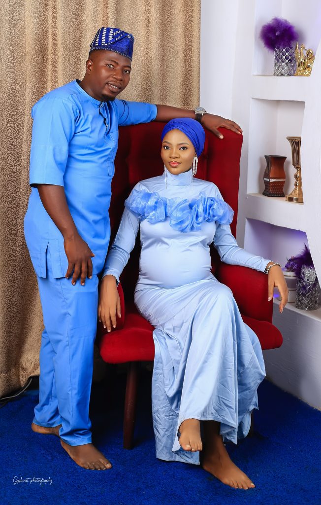 Khair in a sitting position on a blue dress and a blue scarf, with a subtle make up and a dangling earrings. Her cute third trimester baby bump is showing signifying a maternity shoot, her husband is right beside her in a blue buba and Sokoto. 