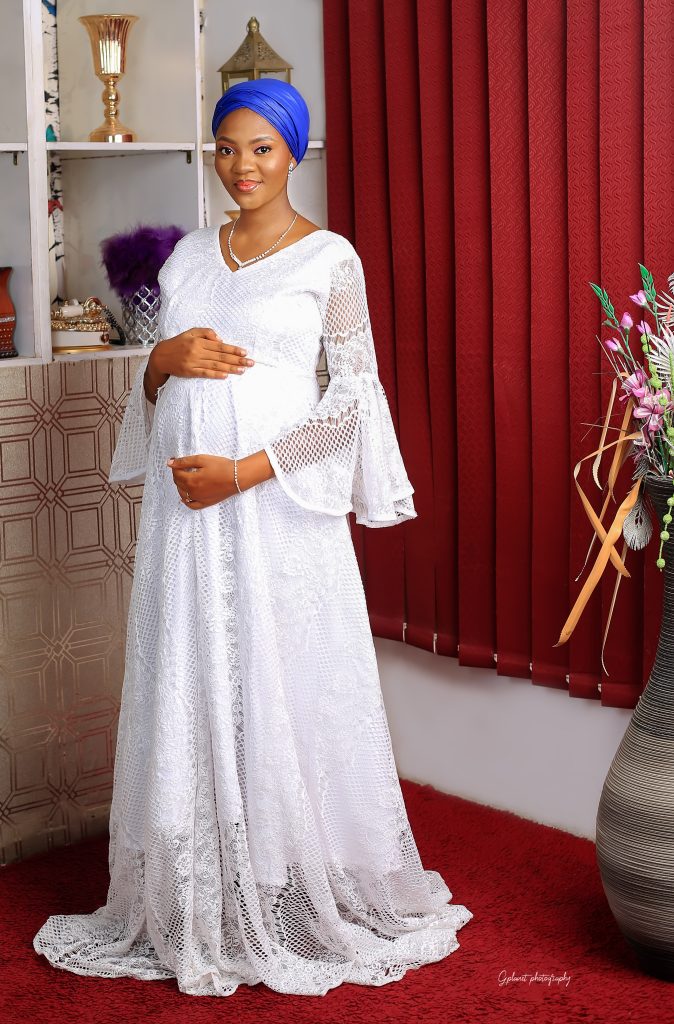 Khair in a standing position on a white dress and a blue scarf, with a subtle make up and a dangling earrings. Her cute third trimester baby bump is showing signifying a maternity shoot.